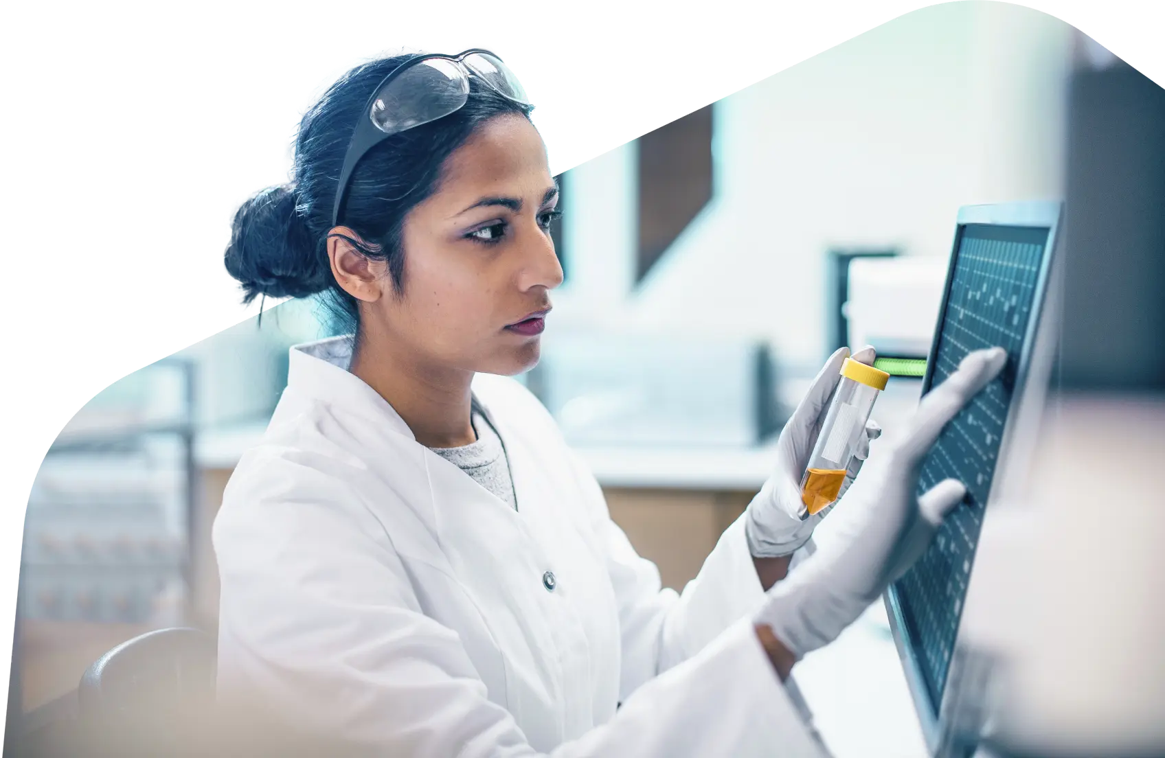 A scientist holds a vial of liquid with one hand while using a touch screen monitor with another.