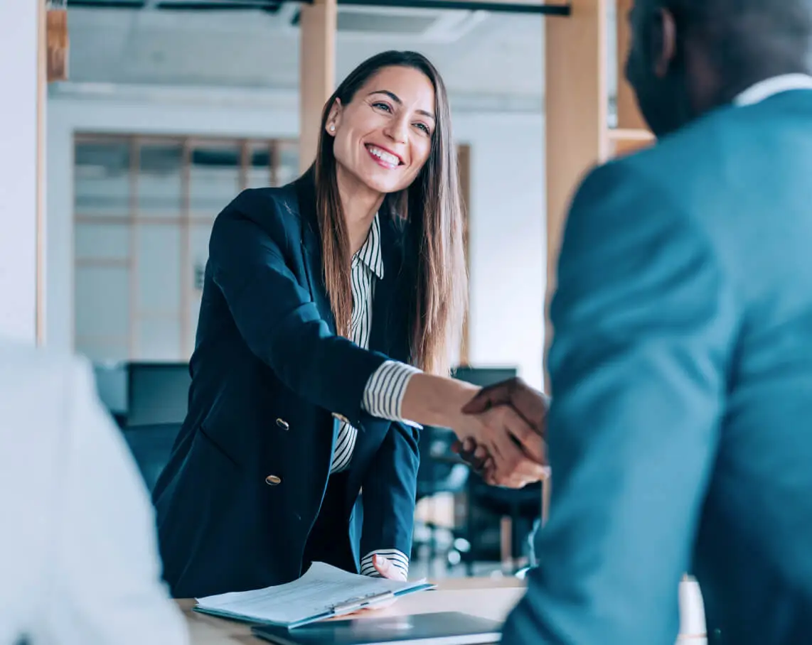 A smiling woman reaches across a desk to shake someone's hand.